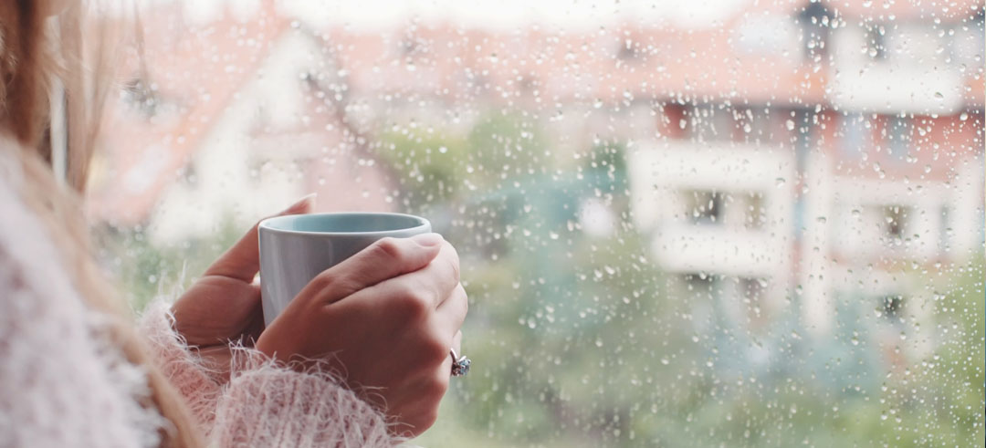 Mujer sosteniendo una taza con ambas manos mientras mira por la ventana en un día lluvioso